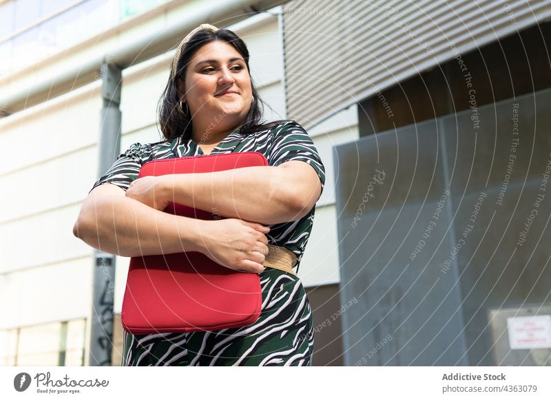 Glückliche Frau mit Laptop-Tasche in der Nähe eines städtischen Gebäudes Lächeln Fall urban modern kurvenreich Stil Farbe Übergröße Streifen Geometrie jung