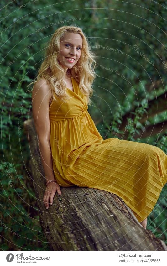 Attraktive blonde Frau in einem stilvollen gelben Kleid posiert auf einem alten Baumstamm gegen Grün im Sommer Blick auf die Kamera mit einem Lächeln in einer Nahaufnahme Porträt