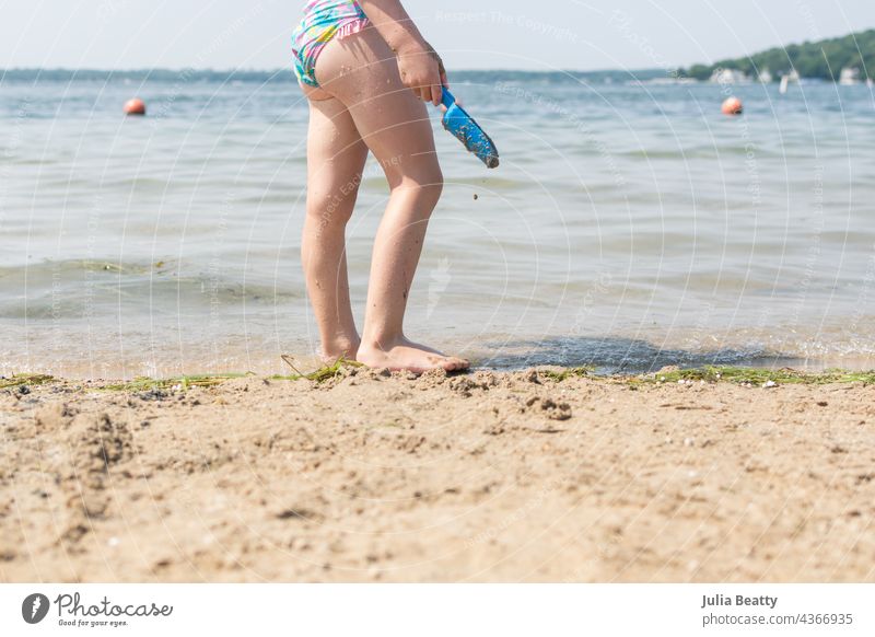 Kleines Mädchen, das am Ufer steht und eine blaue Plastikschaufel in der Hand hält; Spielen am See im Sommer Kind Badeanzug schaufeln Kunststoff Baggerlöffel