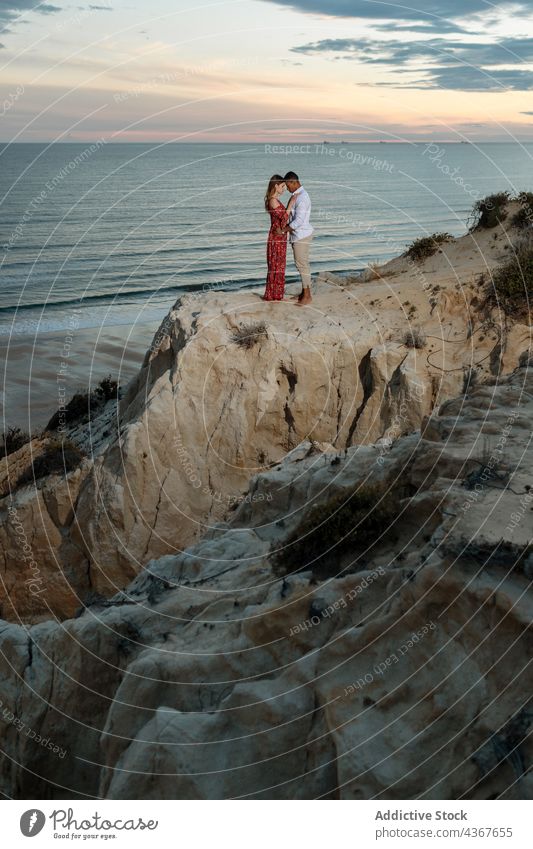 Sanftes multiethnisches Paar steht auf einem Hügel in der Nähe des Meeres bei Sonnenuntergang Umarmen MEER Sommer elegant romantisch Liebe Zusammensein Umarmung