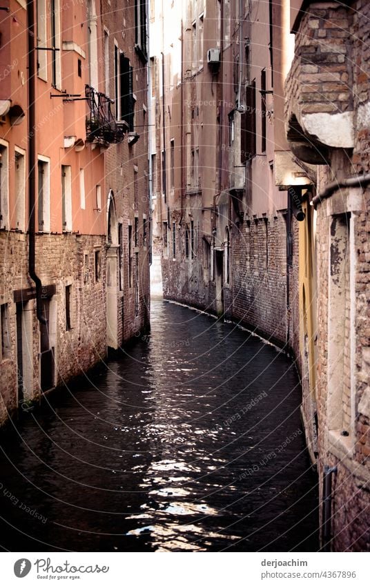 In einer Wasserstraße von Venedig. Alte nicht verputzte  Häuser, die ganz dicht zusammen stehen.Das Wasser schimmert.  Das Sonnenlicht durchdringt das ganze etwas. Der alte Glanz der Häuser ist schon lange verloren  gegangen.