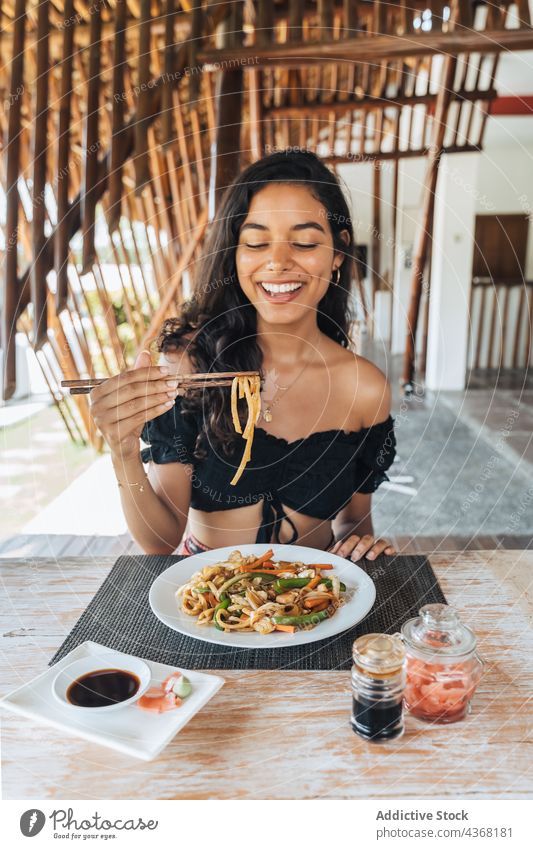 Lächelnder Reisender mit Nudeln zwischen Stäbchen im Restaurant Asiatische Küche Essstäbchen heiter Mittagessen Mahlzeit Tisch Frau genießen Lebensmittel-Stick