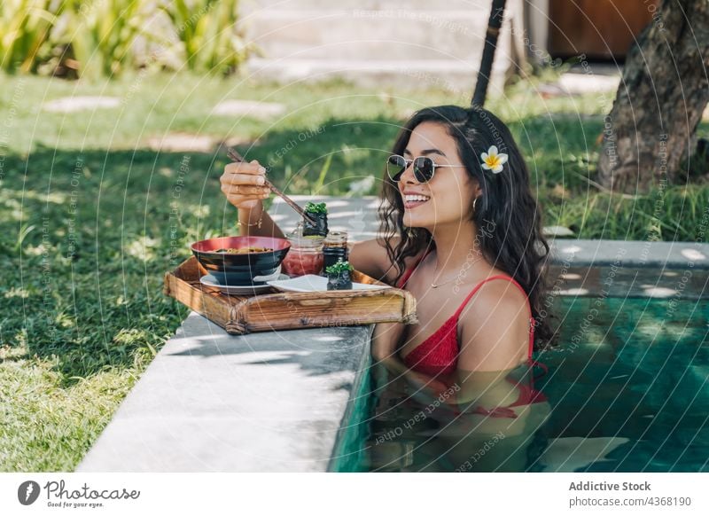 Lächelnder Tourist mit leckeren Nudeln im Pool während eines Ausflugs Asiatische Küche Sushi Resort Frau Essstäbchen Badebekleidung Reisender Gunkan Mittagessen