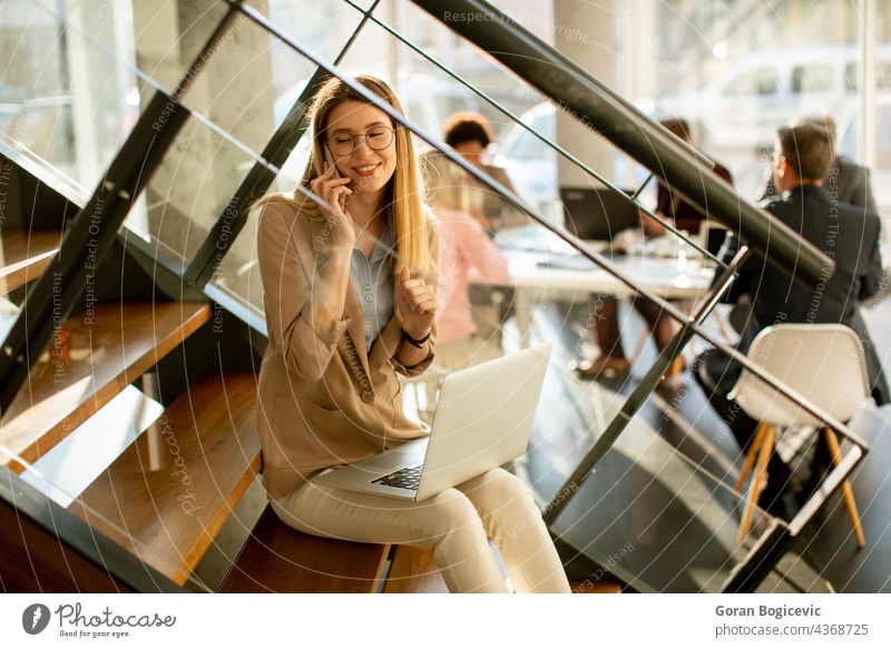 Junge Frau sitzt auf der Treppe mit einem Laptop und benutzt ein Mobiltelefon in einem modernen Büro vor ihrem Team Erwachsener attraktiv schön Business