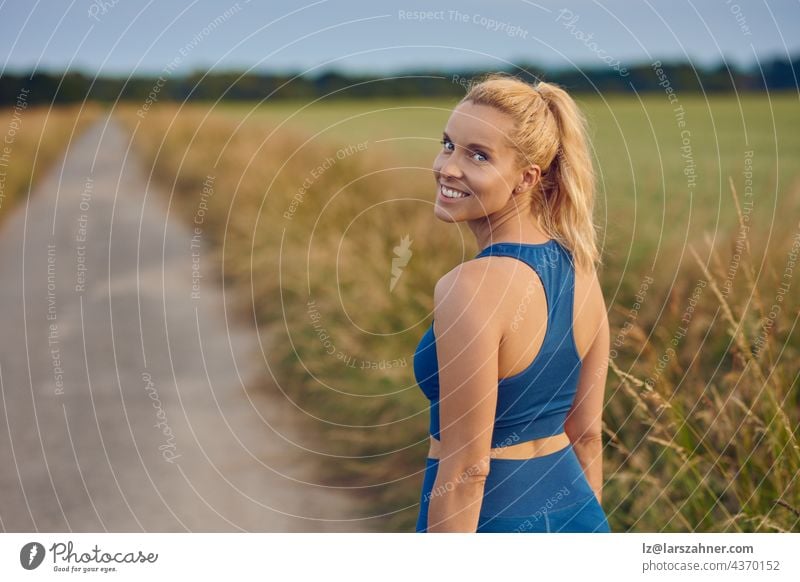Attraktive, fitte, gesunde Frau, die sich umdreht, um in die Kamera zu lächeln, während sie sich darauf vorbereitet, auf einem ländlichen Wanderweg zu joggen, in einem gesunden, aktiven Lebensstil im Freien