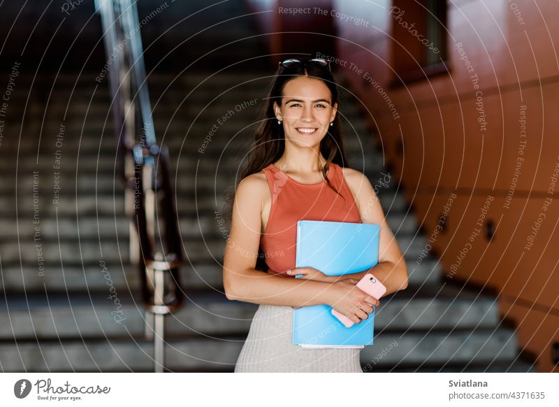 Eine junge Studentin in Freizeitkleidung mit einem Aufsatz in einer Mappe und einem Telefon in der Hand Mädchen Schüler Brille Treppe Frau lässig Smartphone