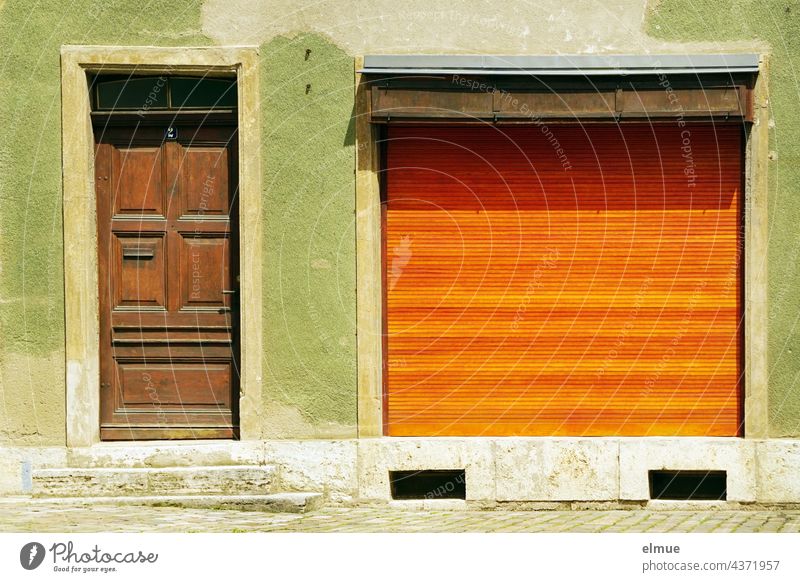 grüne Hausfassade mit brauner Holztür und großem Schaufenster mit einem geschlossenen,  orangefarbenen Rolladen / Betriebsferien / Urlaub Fassade Straße
