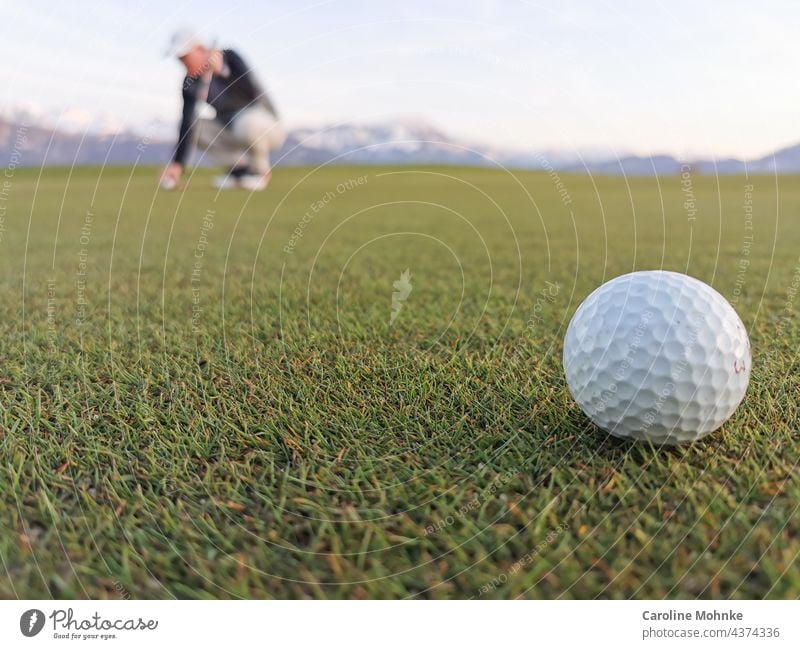 Golfball auf einem Green - Im Hintergrund ein Golfer, der vor einem Alpenpanorama seinen Ball ausrichtet Natur Sport Golfplatz Freizeit & Hobby Spielen