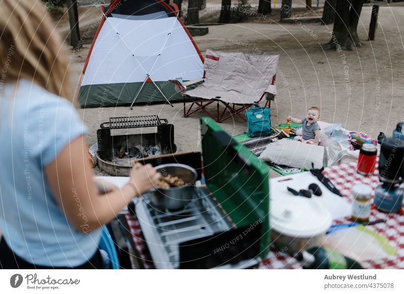 Baby auf dem Boden eines Zeltplatzes sitzend, während die Mutter auf einem Campingkocher kocht blond Kalifornien Lager Wohnmobil Campingplatz Kaukasier Stuhl