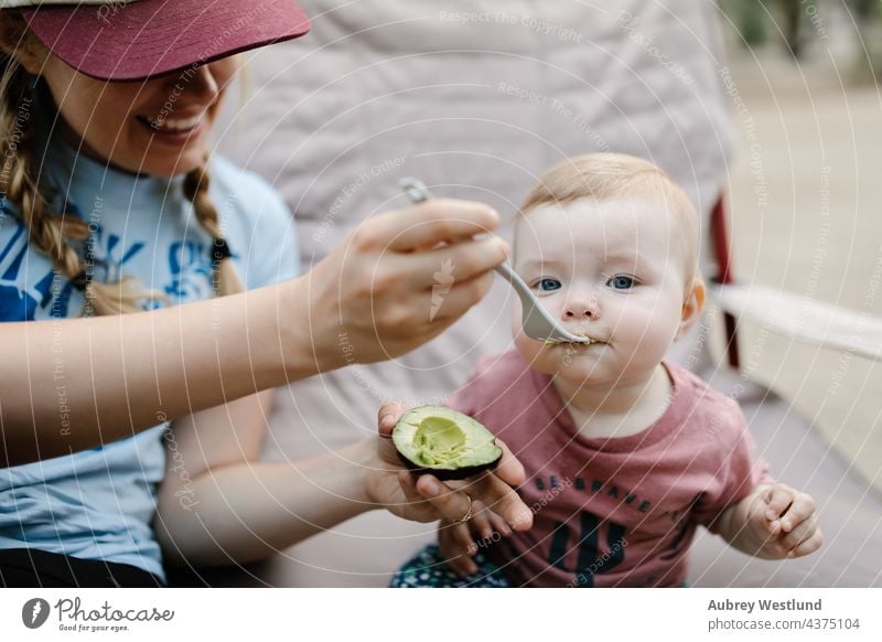 Mutter füttert ihre kleine Tochter mit einer Avocado beim Camping im Freien Baby blond blaue Augen Kalifornien Lager Wohnmobil Campingplatz Kaukasier Stuhl Kind