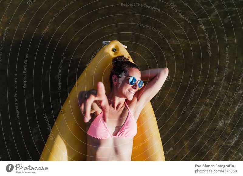 Draufsicht auf glückliche kaukasische Frau, die auf einem gelben Kanu im See liegt, während eines sonnigen Tages, der mit dem Finger auf die Kamera zeigt. Sport, Abenteuer und Natur. Frau in Badehose und Sonnenbrille