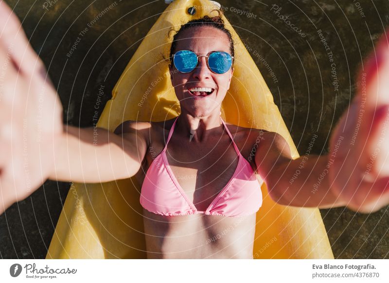 Draufsicht auf glückliche kaukasische Frau, die auf einem gelben Kanu in einem See liegt und an einem sonnigen Tag eine Kamera hält, um ein Selfie zu machen. Sport, Abenteuer und Natur. Frau in Badekleidung und Sonnenbrille