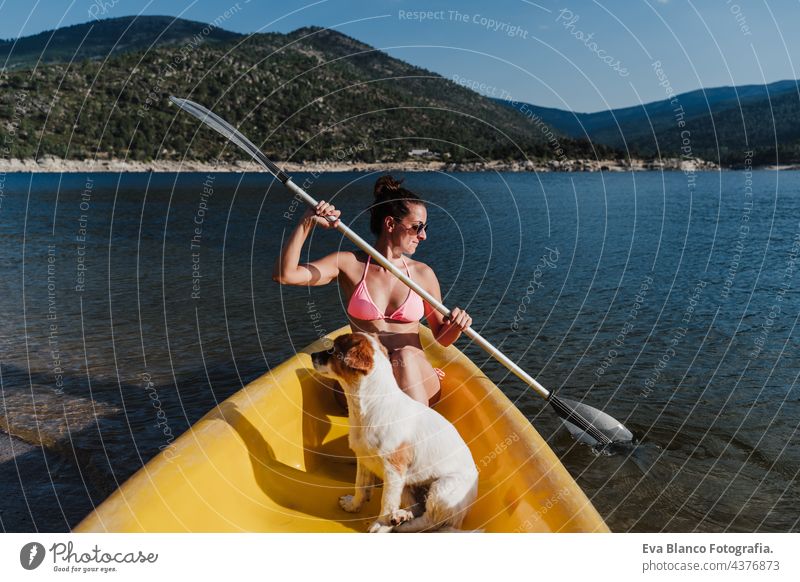 Junge kaukasische Frau und Jack-Russell-Hund sitzen auf gelbem Kanu in See während sonnigen Tag. Frau rudert. Sommerzeit. Haustiere, Abenteuer und Natur