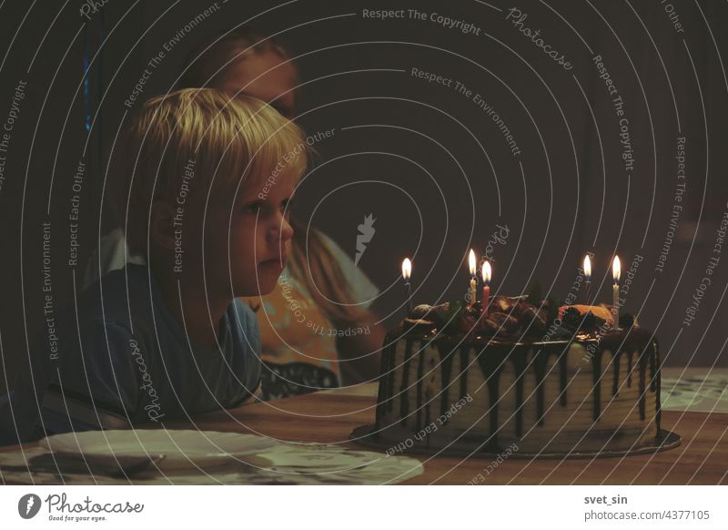 Ein blonder Junge sitzt an einem Tisch in einem dunklen Raum und schaut auf einen Geburtstagskuchen mit brennenden Kerzen. Die Geschwister warten auf der Geburtstagsfeier auf den Nachtisch.