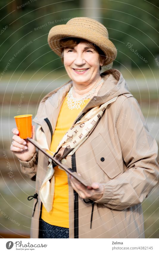 Lächelnde ältere Frau mit Hut, Tablet und einem Glas Kaffee auf Reisen während der Covid-19-Pandemie Senior alt Menschen Gesicht Porträt Sommer Feiertag aktiv
