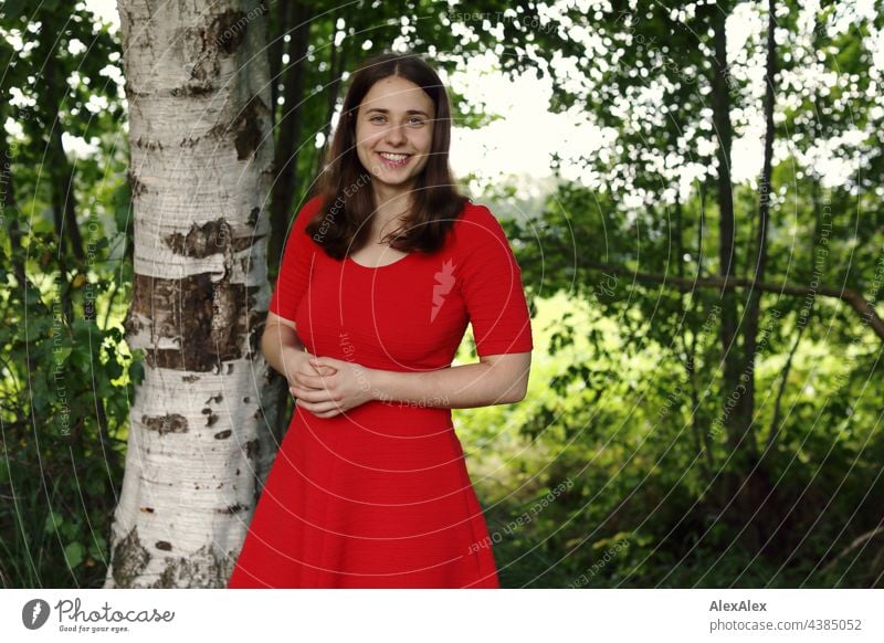 Junge Frau in rotem Sommerkleid steht in der Natur im Grünen und schaut lächelnd in die Kamera sehen grün brünett Kleid rotes Kleid Gras Schönheit und Schönheit
