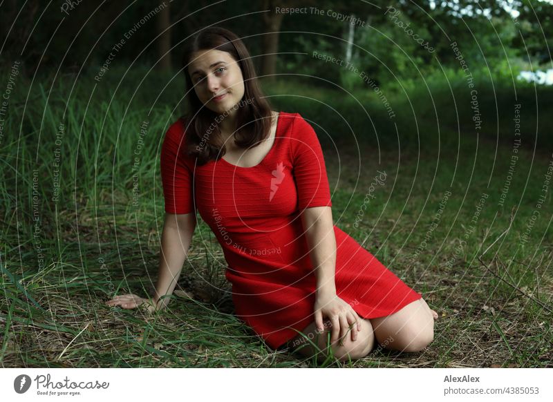 Junge Frau in rotem Sommerkleid sitzt in der Natur im Grünen an einem See und schaut in die Kamera sehen brünett Kleid rotes Kleid Gras Schönheit sportlich