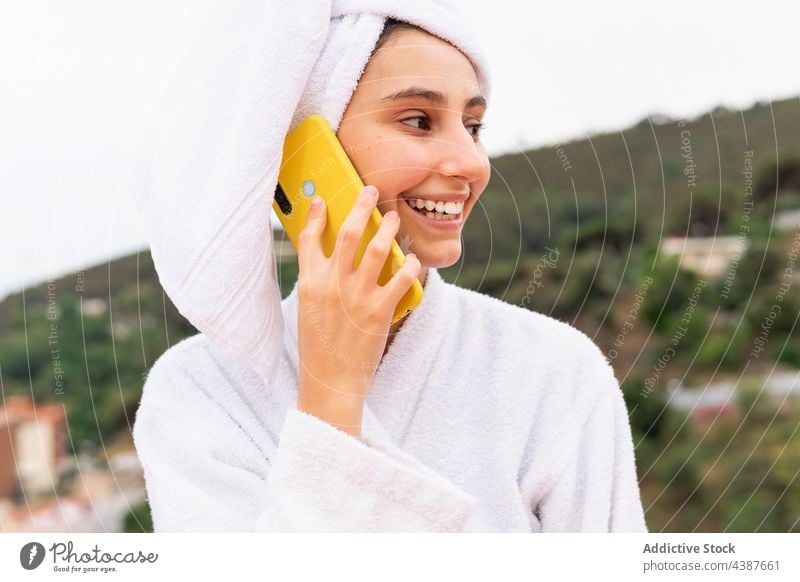 Frau benutzt Smartphone auf Balkon nach Dusche benutzend Hautpflege ruhen Sauberkeit Lächeln soziale Netzwerke Wochenende Browsen Handtuch Bademantel