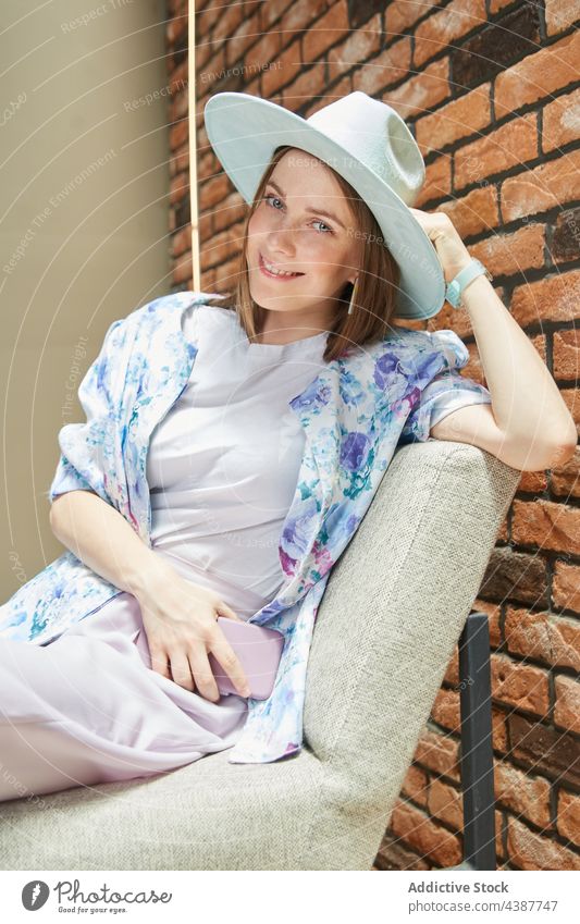 Trendige Frau mit Hut, die sich auf einem Stuhl an einer Backsteinmauer ausruht Lächeln Stil herzlich freundlich Art Backsteinwand Porträt Smartphone Apparatur