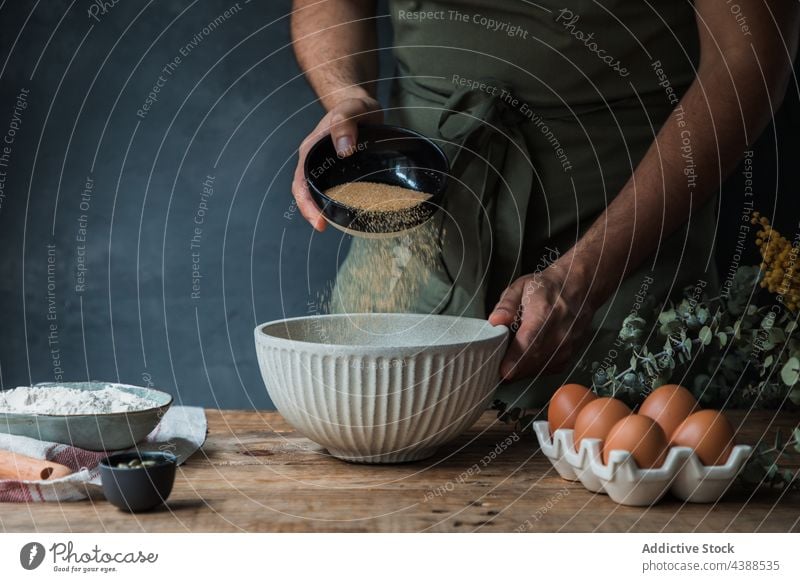 Kropfenkoch gibt Rohrzucker zum Teig Mann Koch Gebäck Brot hinzufügen Schalen & Schüsseln Bestandteil rustikal Tisch männlich Schürze Ei Mehl Lebensmittel