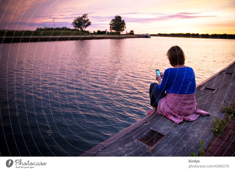 Frau sitzt bei Sonnenuntergang auf einer Kaimauer und schaut auf ihr Smartphone Wasser Landschaft Horizont Sonnenlicht Freizeit sitzen geniesen Dämmerung Fluss