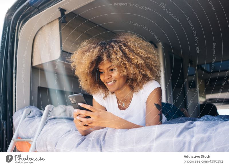Fröhliche schwarze Frau mit Smartphone auf dem Bett eines Wohnmobils Wohnwagen benutzend Autoreise Lächeln ruhen Wochenende Sommer Urlaub jung Browsen heiter