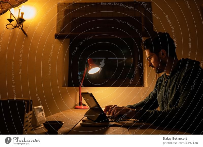 Junger Mann arbeitet nachts mit seinem Tablet in der Dämmerung Nacht Telearbeit Tablette Licht Dunkelheit Konzentration Arbeit Brille Schreibtisch Internet