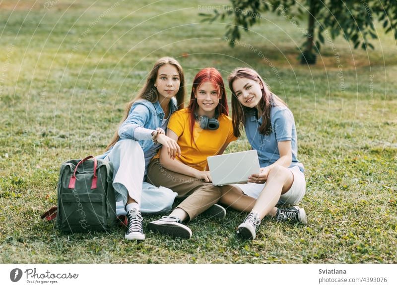 Teenager-Mädchen sitzen mit einem Rucksack und einem digitalen Tablet auf einer grünen Wiese in einem Park und bereiten ein gemeinsames Projekt vor. Bildung und Wissen, Lebensstil von Teenagern