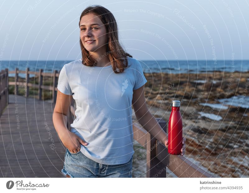 Teenager-Mädchen im T-Shirt steht und hält wiederverwendbare Wasserflasche in den Händen Flasche MEER Sonnenuntergang Attrappe Beteiligung rot blau hell-blau