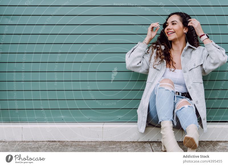 Glückliche Frau, die in der Nähe einer Hauswand in der Stadt sitzt Zahnfarbenes Lächeln charmant Stil Großstadt Straße urban trendy Mode Vorschein Gebäude Wand