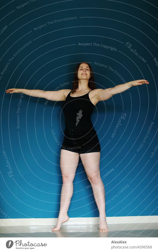 Junge Frau in schwarzem Trägertop und Hotpants steht barfuß vor einer petrolblauen Wand und hebt die Arme in einer Tanzpose brünett sportlich 18-30 Jahre