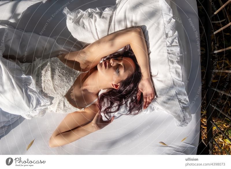 Porträt einer attraktiven, zufriedenen, jungen, sexy, dunkelbraunhaarigen Frau, die entspannt im Bett liegt und sich mit frischen, weichen, weißen Laken und Kissen in der Morgensonne kuschelt, Kopierraum