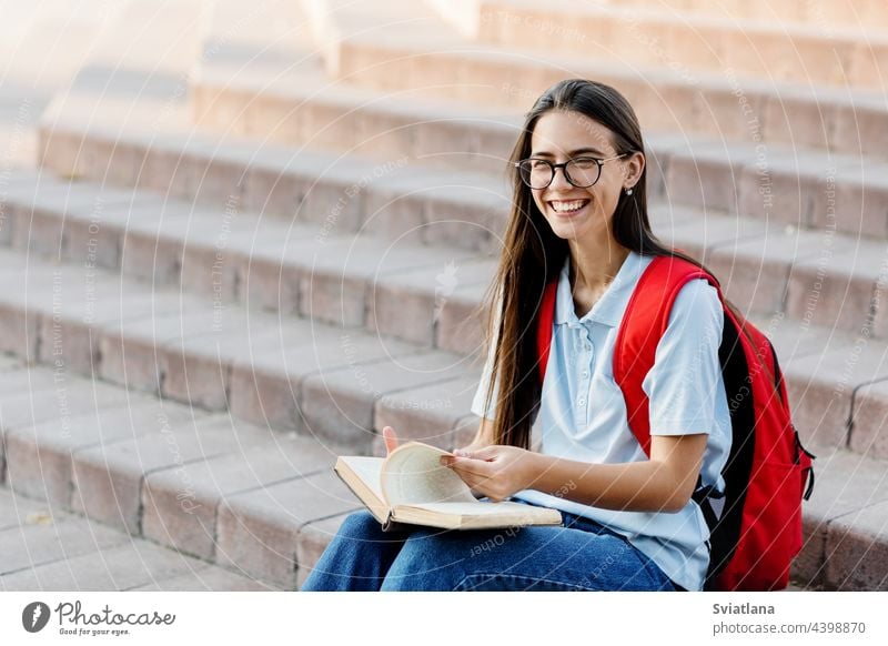 Eine hübsche Studentin mit Brille und Rucksack sitzt auf einer Treppe in der Nähe der Universität und liest ein Buch, um sich auf Vorlesungen oder Prüfungen vorzubereiten. Ausbildung und Bildung