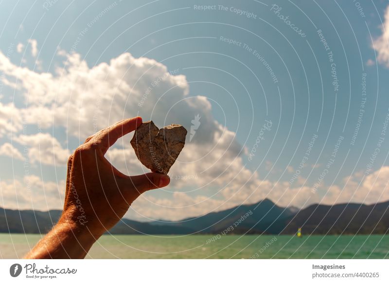 Hand, die ein Herz hält. Steinherz in einer weiblichen Hand auf dem Hintergrund des Himmels und des Sees. Liebe zur Natur. Voralpen-Berge und Walchensee in Bayern, Deutschland