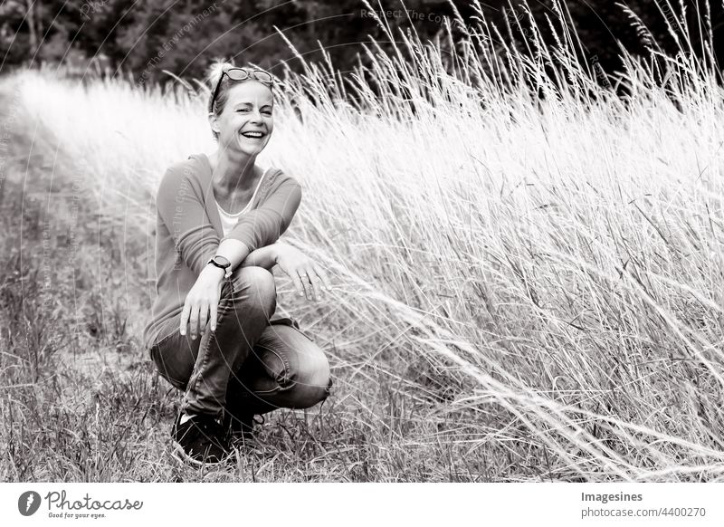 Schwarz-Weiß-Porträt einer lächelnden glücklichen und charmanten Frau in die Kamera schaut. Frauenporträt im Sommer hockend auf einer Wiese. Gras Erwachsene