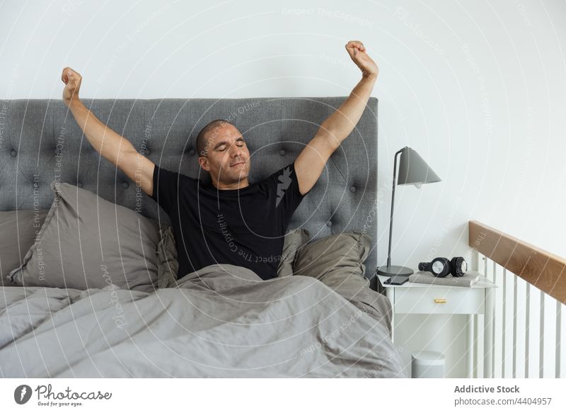 Zufriedener Mann, der sich nach dem Aufwachen auf dem Bett streckt Dehnung Morgen Schlafzimmer sorgenfrei früh aufwachen Komfort männlich genießen sitzen weich