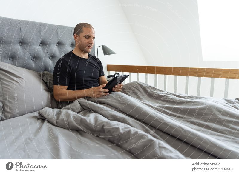 Mann surft morgens im Bett auf seinem Tablet Browsen Tablette Morgen wach Schlafzimmer benutzend Surfen online männlich heimwärts Apparatur Gerät Komfort