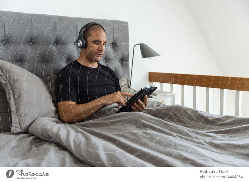 Mann benutzt morgens zu Hause im Bett ein Tablet Morgen Browsen benutzend Tablette zuhören Musik Schlafzimmer wach männlich Apparatur ruhen Gerät