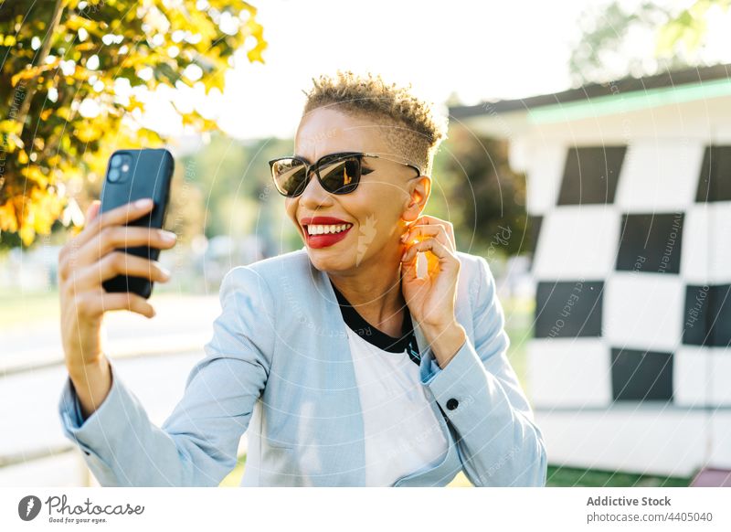 Fröhliche schwarze Frau mit Smartphone bei einem Videogespräch Videoanruf reden heiter Mode Sonnenbrille Freizeit Park benutzend Apparatur Internet Gerät
