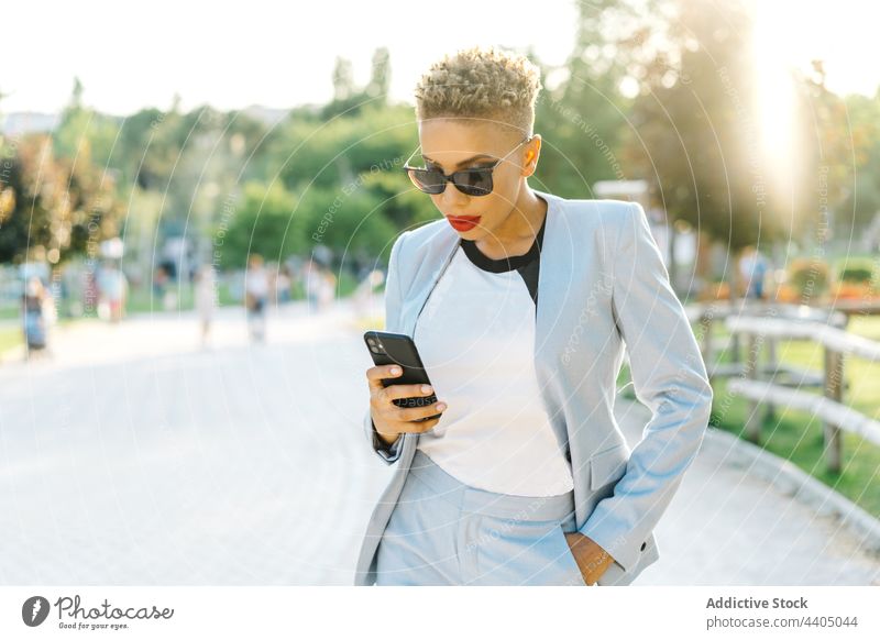 Trendige schwarze Frau, die beim Gehen auf dem Bürgersteig mit ihrem Smartphone chattet plaudernd Mode Stil Individualität Sonnenbrille Spaziergang Straßenbelag