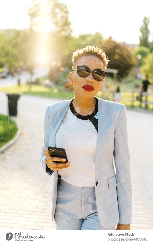 Trendige schwarze Frau, die beim Gehen auf dem Bürgersteig mit ihrem Smartphone chattet Porträt Mode Stil Individualität Sonnenbrille Spaziergang Straßenbelag