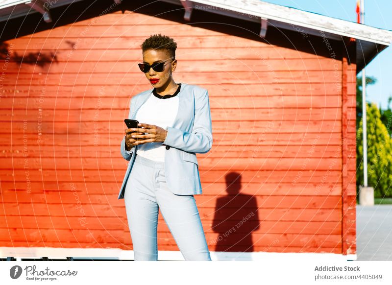 Stilvolle schwarze Frau mit Sonnenbrille und Smartphone auf der Straße Telefon Mode stylisch Individualität cool benutzend Apparatur Gerät Straßenbelag