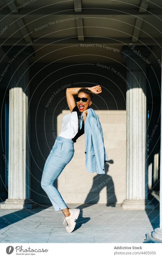Glückliche schwarze Frau in trendiger Kleidung auf Zehenspitzen in der Stadt Mode Stil Spaß haben Individualität Sonnenbrille Energie Straßenbelag stylisch