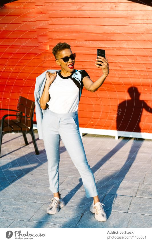 Stylische schwarze Frau mit Sonnenbrille, die auf der Straße mit ihrem Smartphone spricht sprechen Mode stylisch Individualität Hand-in-Tasche cool benutzend