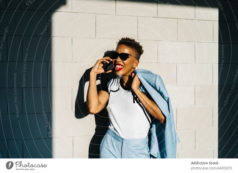 Trendige ethnische Frau mit Sonnenbrille, die auf der Straße mit ihrem Smartphone spricht Mode Stil reden herausstechend Zunge Individualität cool Haarschnitt