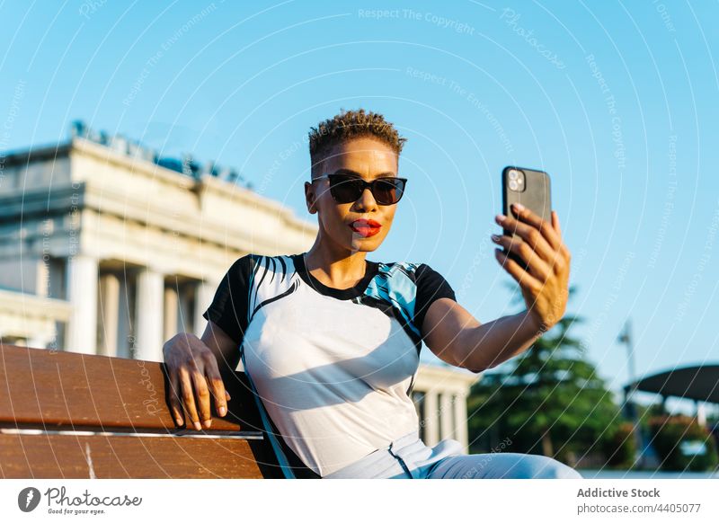 Trendy ethnischen Frau, die Selfie auf Smartphone auf städtischen Bank Mode Stil Sonnenbrille Gedächtnis Moment Stadt benutzend Apparatur Gerät selbstbewusst