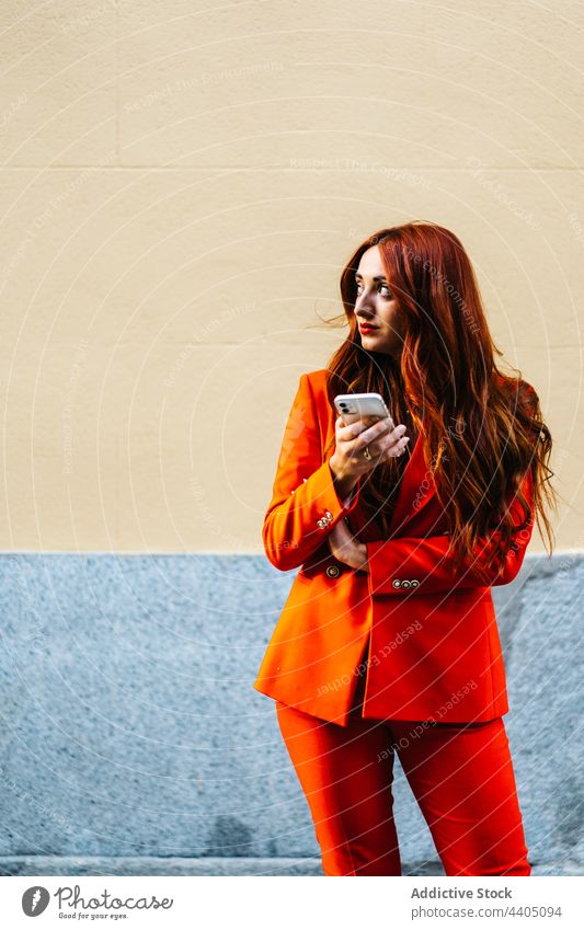 Trendige Frau im orangefarbenen Anzug surft auf ihrem Smartphone benutzend Farbe lebhaft Rotschopf Browsen Surfen Straße Stil Telefon Großstadt Internet Outfit