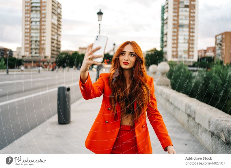 Lächelnde Frau mit Ingwerhaar nimmt Selfie auf der Straße Großstadt Smartphone Selbstportrait rote Haare orange Anzug trendy heiter Stil urban positiv Mode