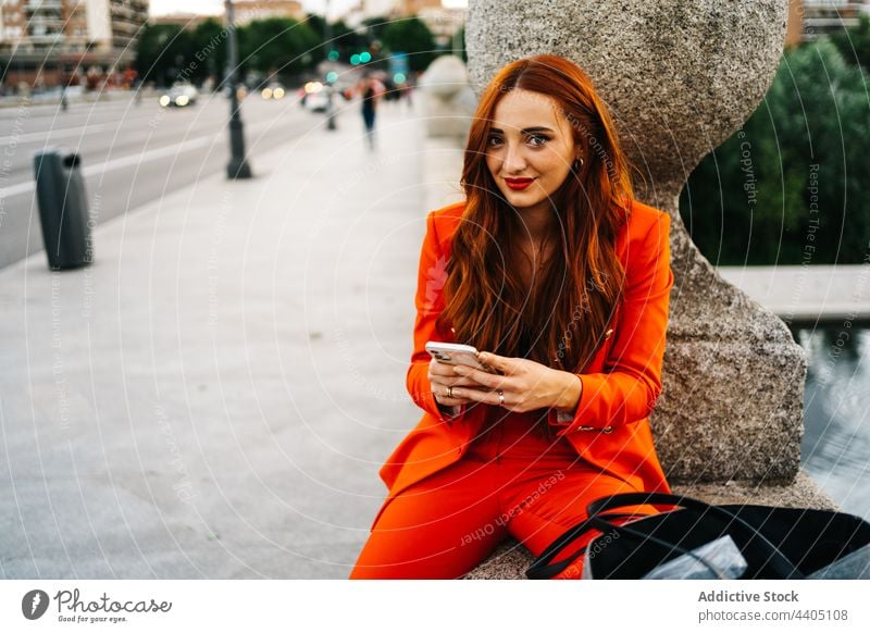 Fröhliche rothaarige Frau in orangefarbenem Anzug, die auf der Straße auf ihrem Smartphone surft Browsen Rotschopf trendy Farbe hell benutzend Apparatur Stil