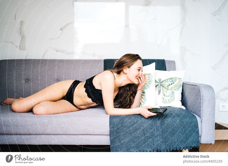 Fröhliche Frau in Unterwäsche, die im Wohnzimmer auf ihrem Smartphone surft Browsen Dessous benutzend Nachricht heiter Sofa heimwärts Mobile soziale Netzwerke